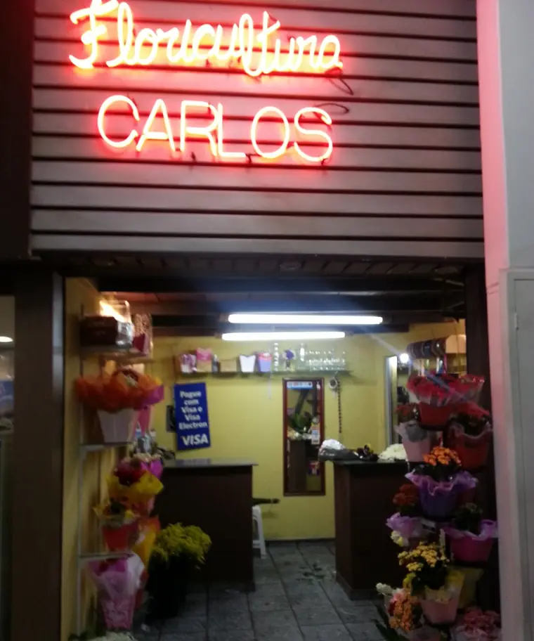 Entrada da loja com o letreiro neon Floricultura Carlos e flores expostas dos dois lados